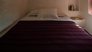 Een slaapkamer met een gebogen plafond, een bed met een rode sprei en een klein plankje met boeken.