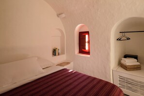Un dormitorio con una cama, un estante pequeño con artículos de aseo, una ventana con marco rojo y un radiador con toallas.