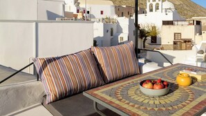 Een terras op het dak met een zachte zithoek, een betegelde tafel met fruit, en uitzicht op witte gebouwen met een klokkentoren.
