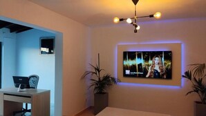 En moderne stue med et fladskærms-tv på væggen, et træskrivebord med en bærbar computer og to potteplanter.