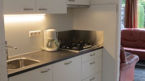 Una cucina compatta con piano cottura a gas, moka e lavello. Si vede una finestra con tende e una sedia rossa.