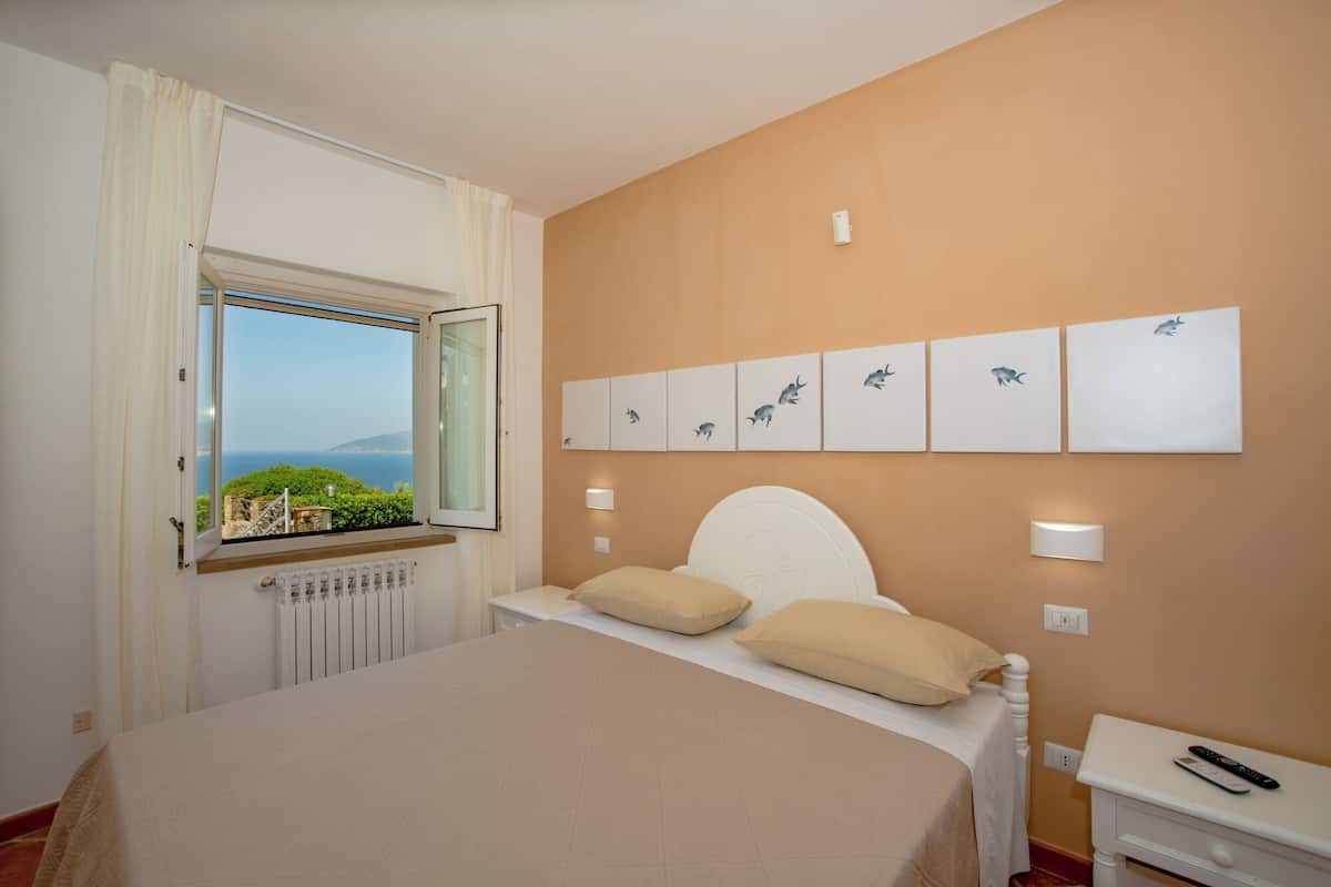 Een slaapkamer met een bed, een raam met gordijnen, een radiator, kunst aan de muur en een nachtkastje met een afstandsbediening.