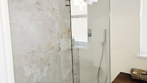 Un espace douche clos par des parois de verre, avec un mur carrelé, une pomme de douche visible et une petite table en bois, à côté de laquelle se trouve un panier près de la fenêtre.