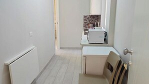 Un couloir étroit avec un radiateur, une porte blanche et une petite table accompagnée d’une chaise.