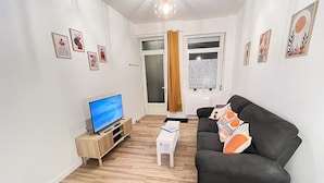 A living room with a black sofa, a wooden TV stand, and a small white table. - Le Duplex 68m2 apartment close to shops, town center and beach (Calais)