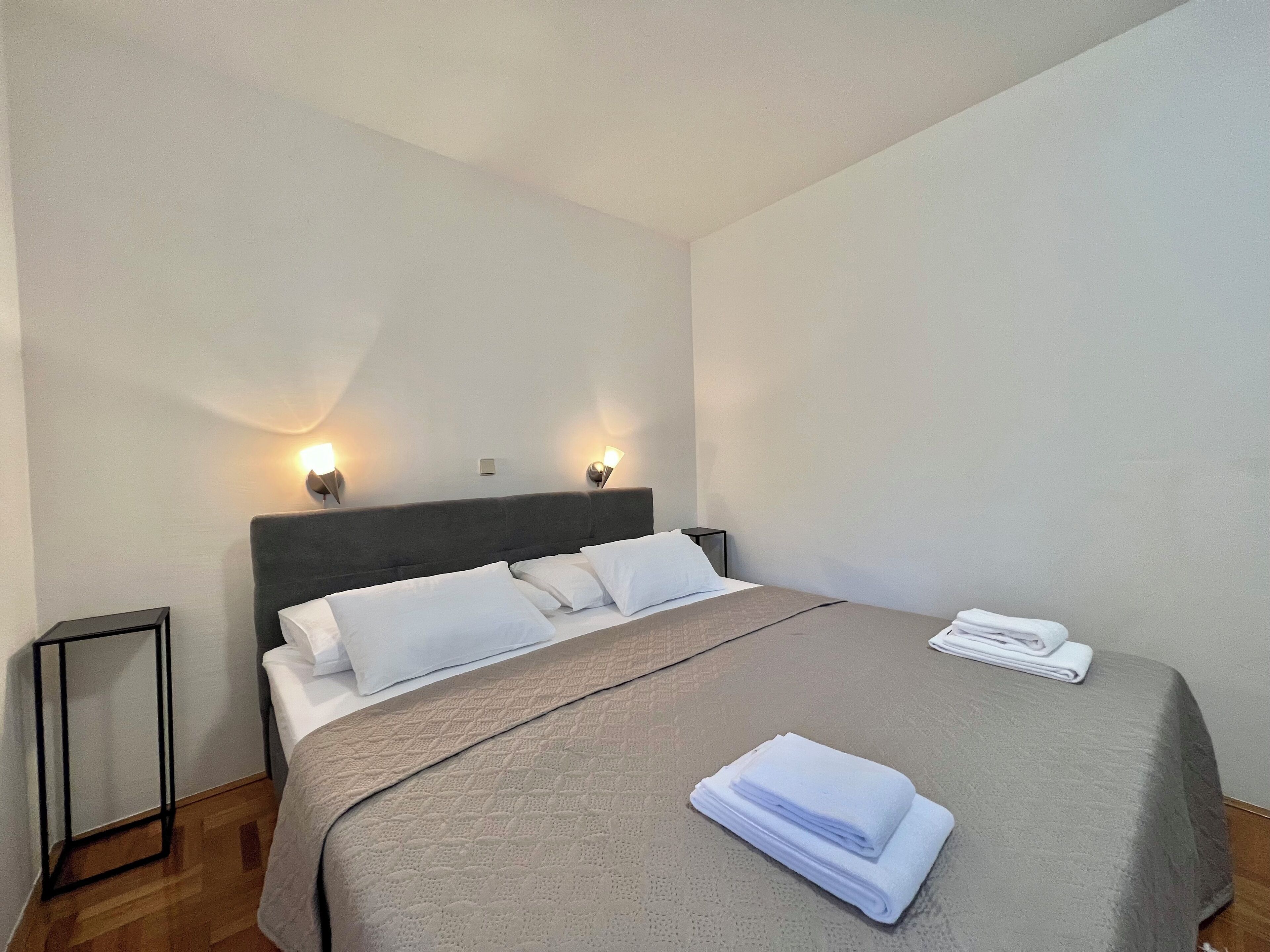 A neatly made bed with a grey bedspread and white pillows, two bedside lamps, and a folded white towel on the bed.
