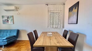 Ein Essbereich mit einem Holztisch und sechs Stühlen, einem blauen Sofa, einem Fenster mit Spitzenvorhängen und einem Bild an der Wand.