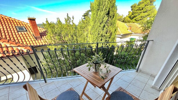 Ein Balkon mit einem Holztisch, zwei Stühlen, einem Blumentopf und Blick auf Bäume und Gebäude.