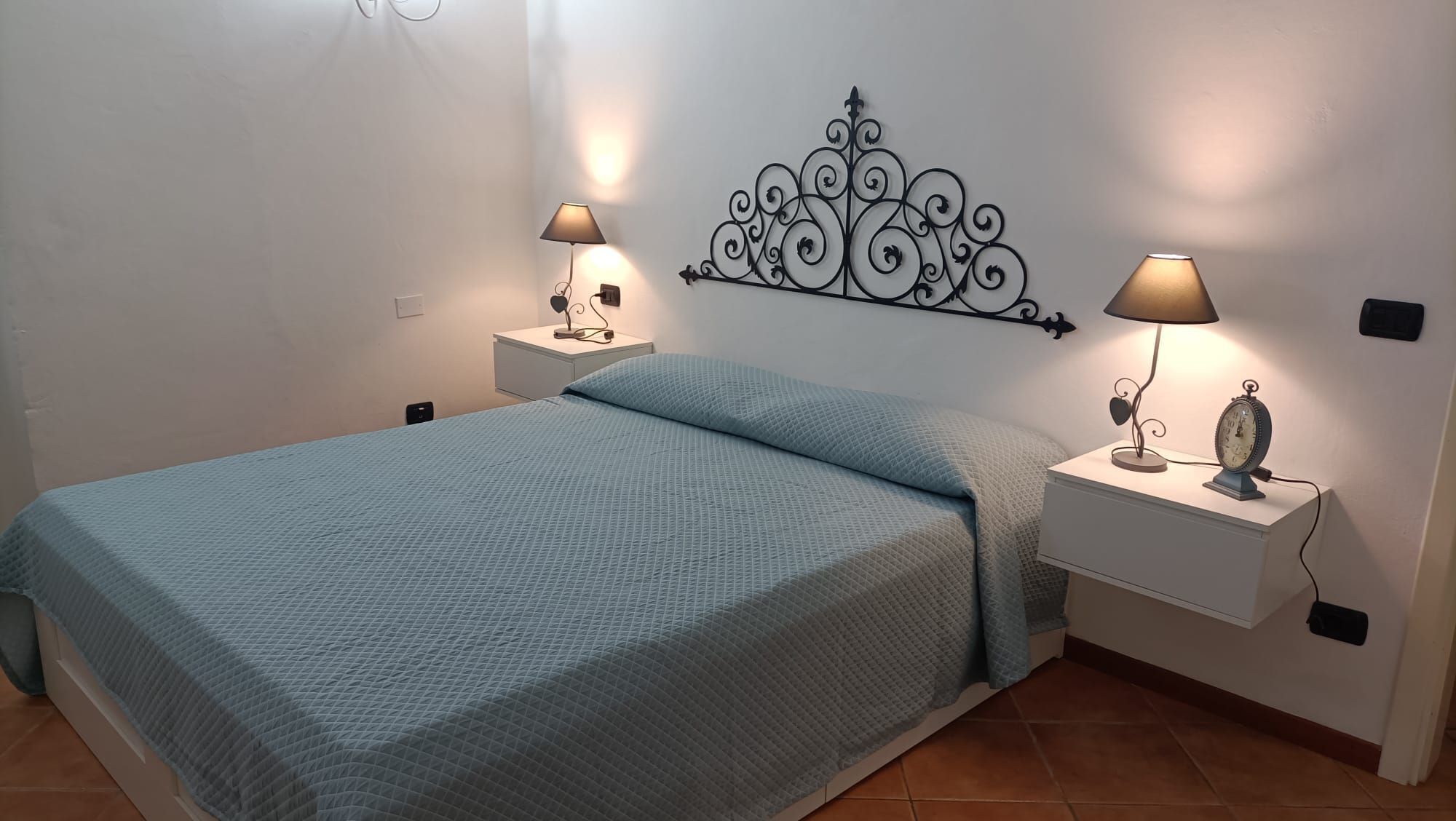 Una camera da letto con un letto, due comodini con lampade, un elemento decorativo a parete e una sveglia.