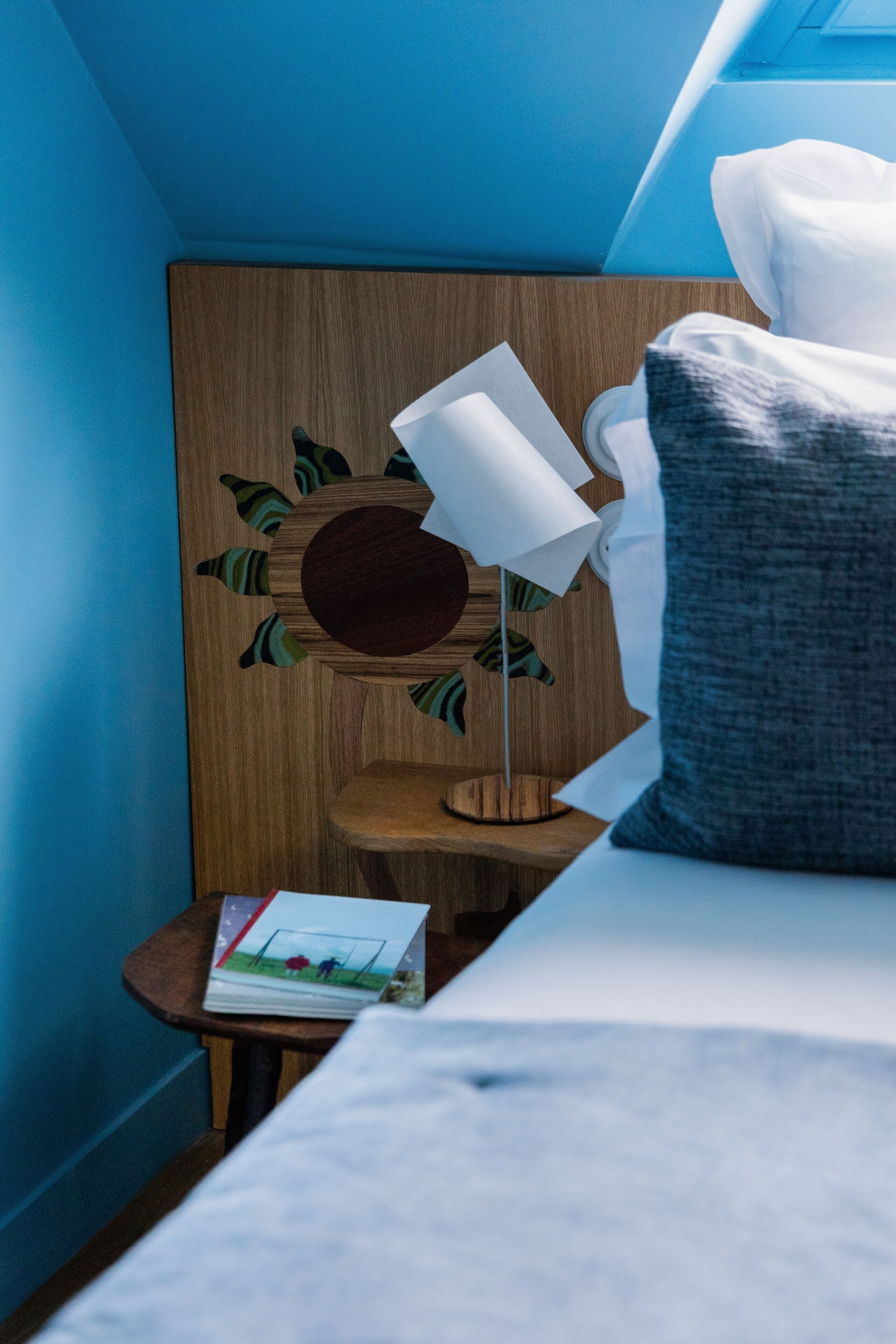 Une chambre à coucher avec un lit, une table de chevet avec une lampe, une tête de lit en bois ornée d’un motif solaire et un oreiller.