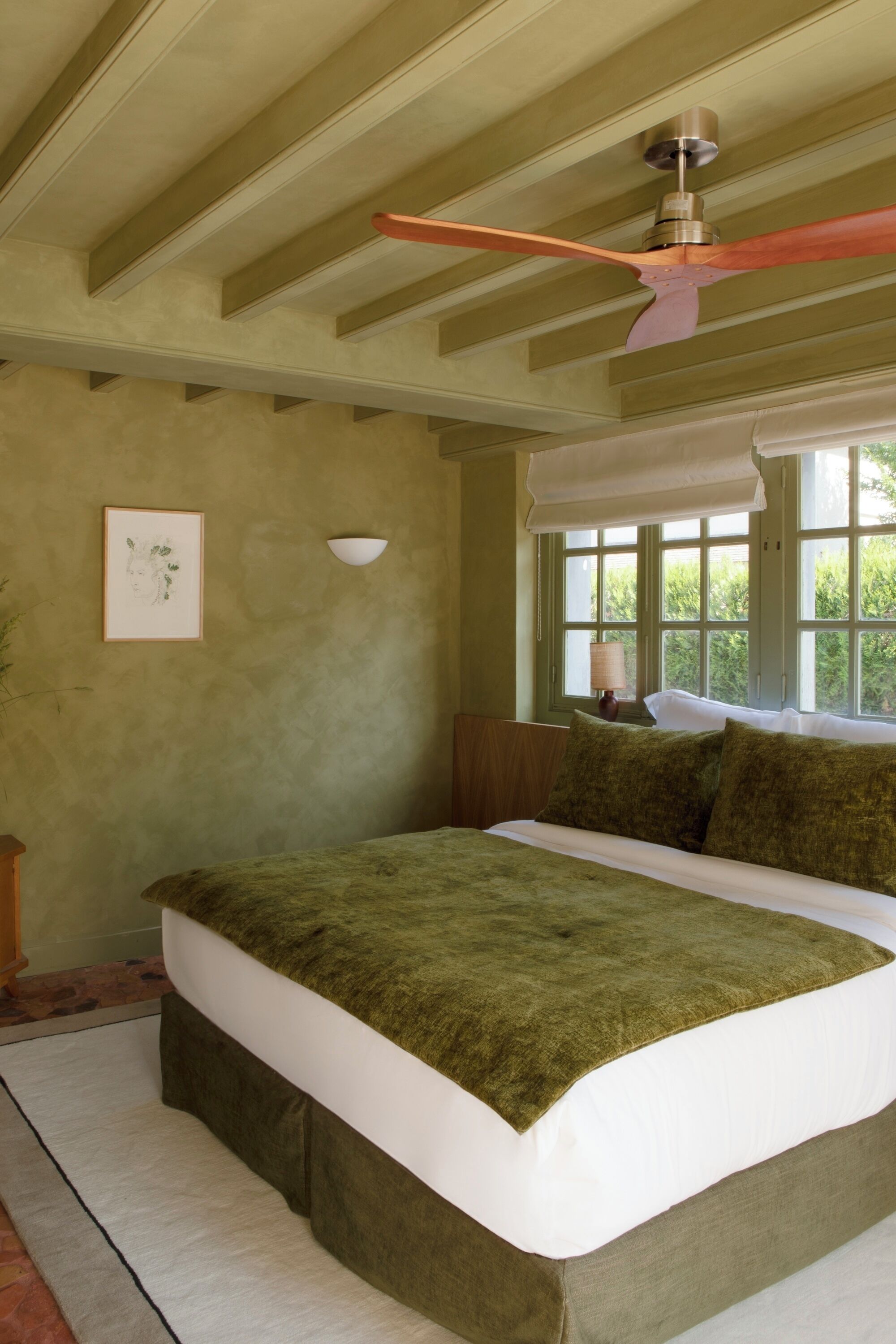 Un dormitorio con una cama grande, un ventilador de techo, una ventana con vistas a zonas verdes y un cuadro en la pared.