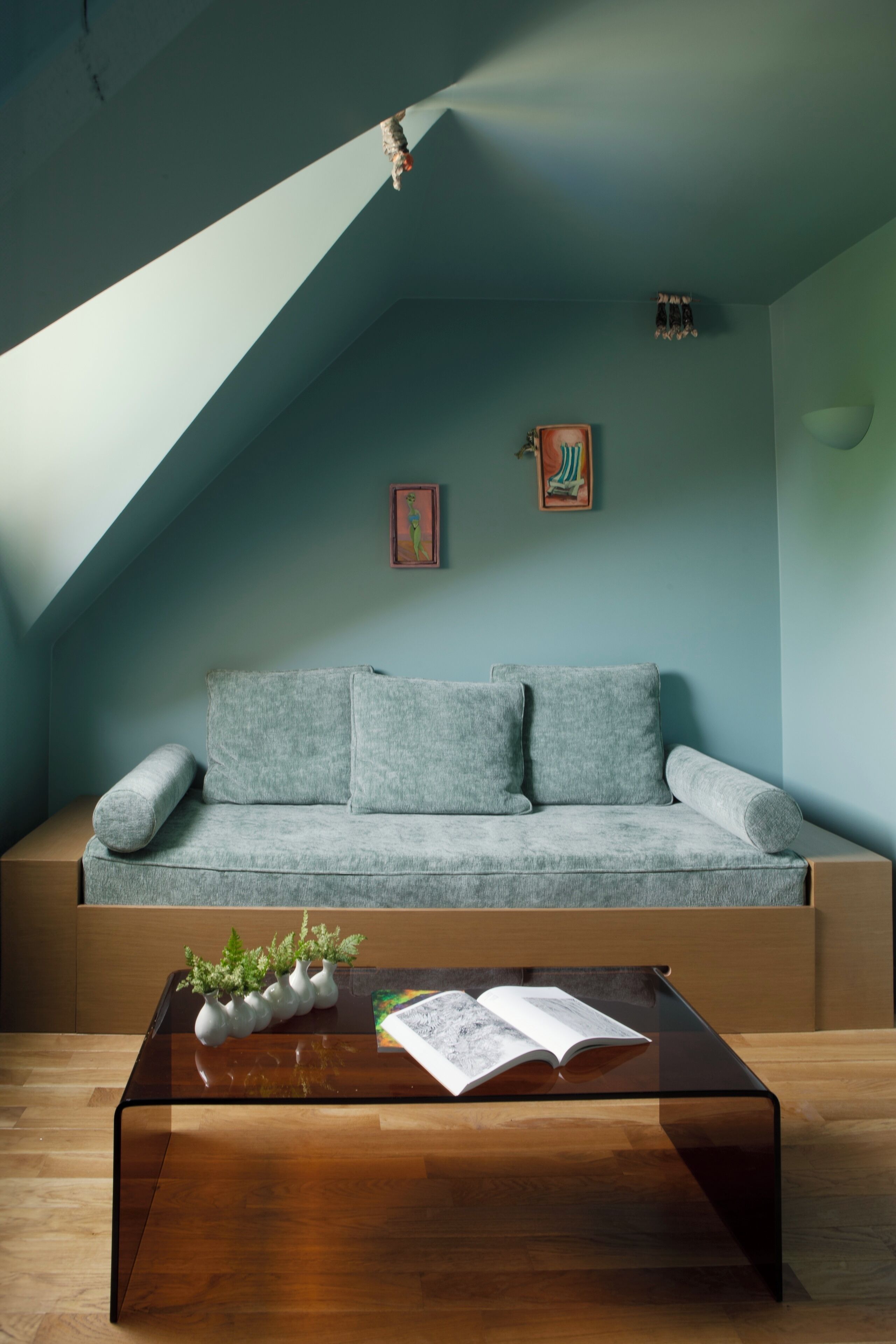 A modern loft bedroom with a sloped ceiling, a grey sofa, a glass coffee table, and two framed artworks.