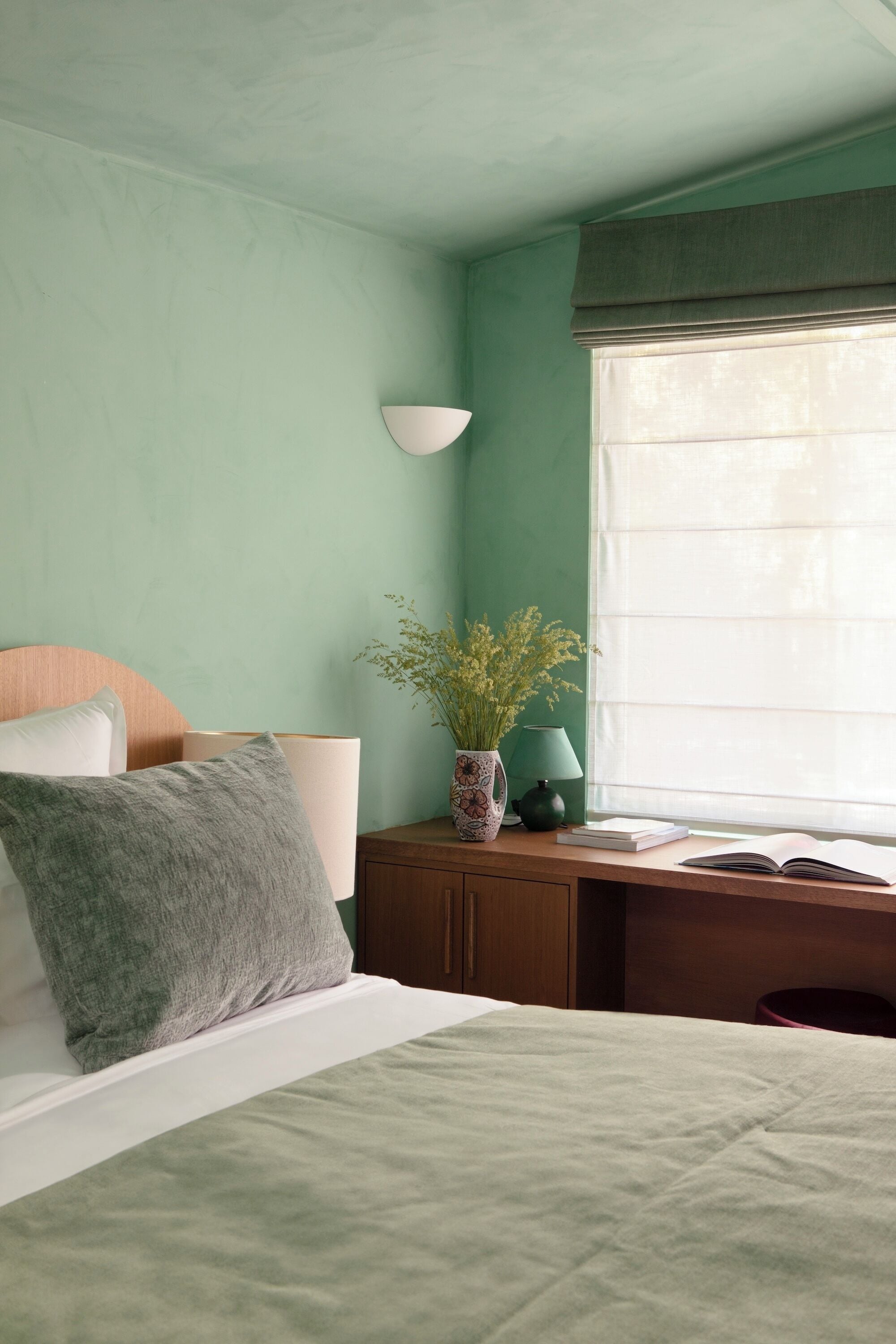 Une chambre avec un lit, une table de chevet avec une lampe, un vase de fleurs et une fenêtre avec des rideaux.