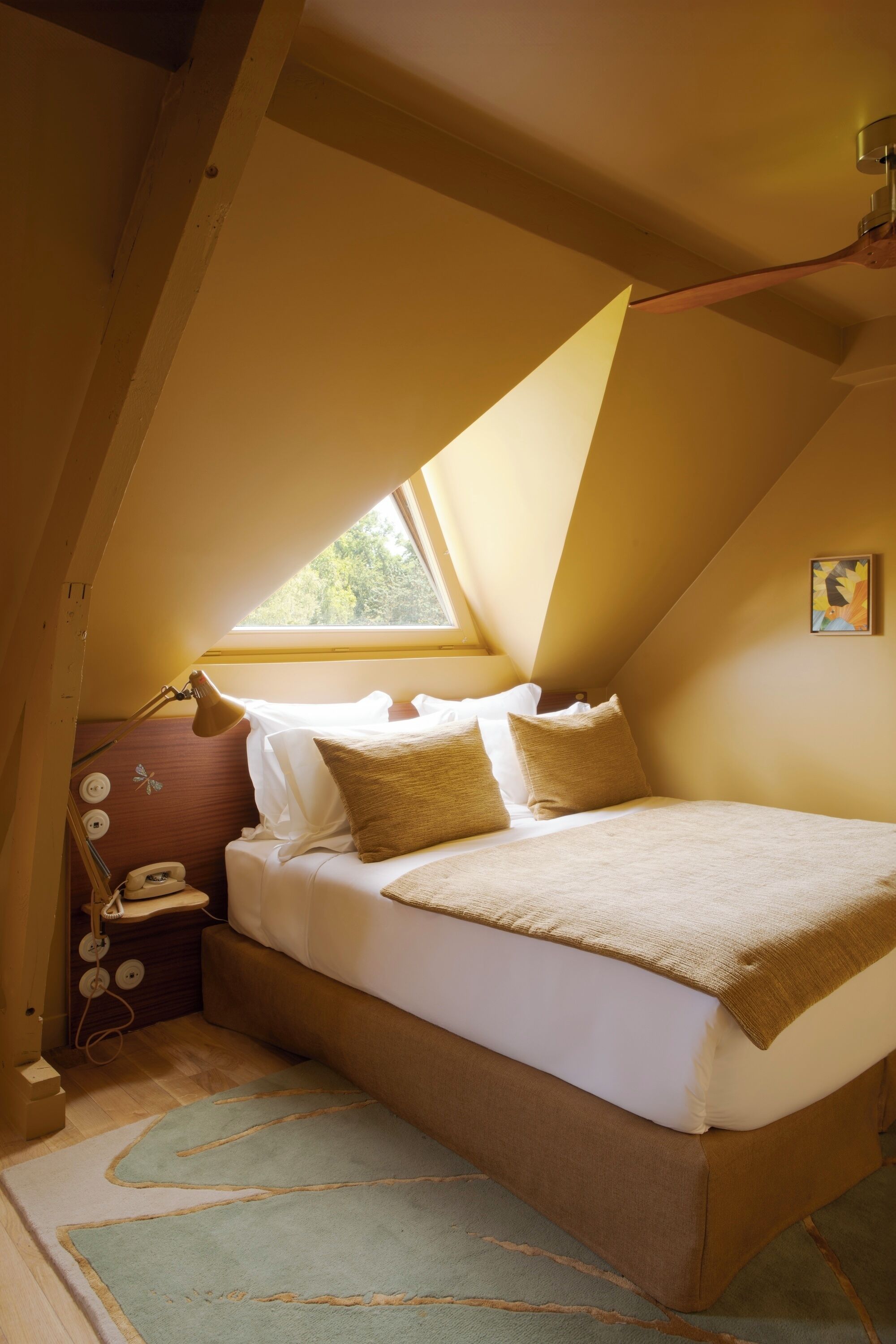 Quarto com teto inclinado, cama com roupa de cama branca, manta marrom e janela que oferece luz natural.