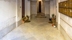 A hallway with marble floors, arched ceilings, and hanging lanterns. There are potted plants along the walls and a staircase in the background.