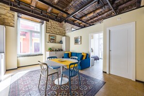 A spacious living area with a high ceiling, a stone wall, a dining table with chairs, a blue sofa with yellow cushions, and a patterned rug.