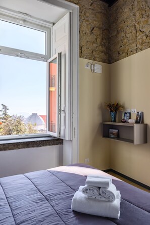 A bedroom with a bed, a window with a view of buildings, a shelf with decorative items, and a wall with a textured stone pattern.