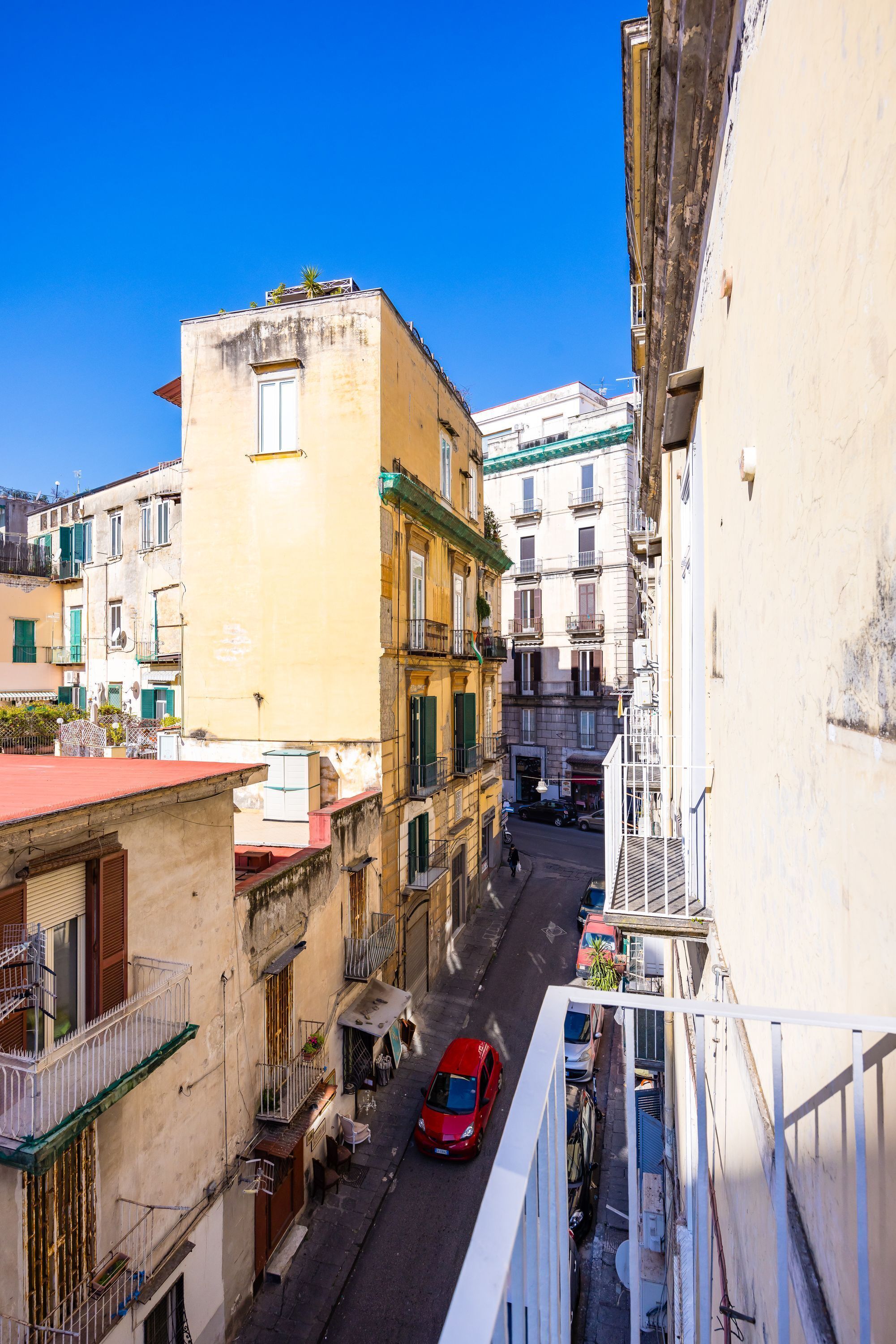 Une rue étroite bordée d’immeubles dotés de balcons, avec une voiture rouge garée sur le côté.