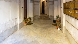 Un couloir doté d’un sol en marbre, de murs blancs et d’une série d’ouvertures en arc menant à différentes pièces. Des luminaires suspendus, de style lanterne, et des plantes en pot décorent les murs.