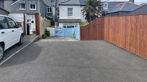 Une allée menant à une résidence, avec une voiture blanche garée sur le côté gauche, un portail bleu et une clôture en bois.