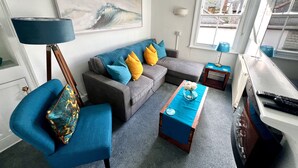 A living room with a grey sofa, blue armchair, and a coffee table with a vase. There is a painting on the wall and a window with a view of buildings.