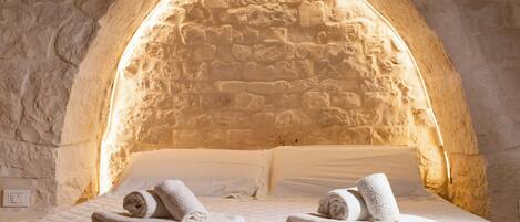 A room with a stone arch ceiling, a bed with white bedding and rolled towels, and a quilted mattress.