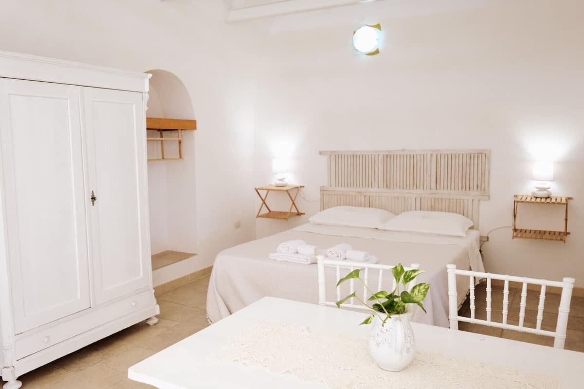 Un dormitorio blanco con cama, mesita de noche, armario y una mesa con una planta.
