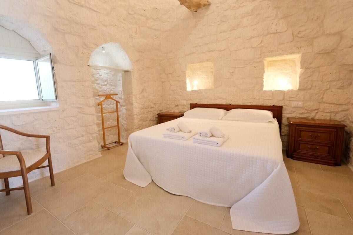 Un dormitorio con pared de piedra, una cama con sábanas blancas, una silla de madera y una mesita de noche.