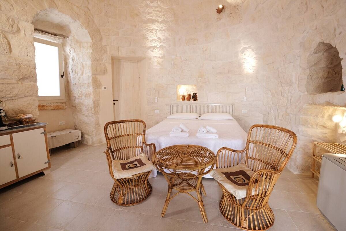 Una habitación acogedora con una cama, dos sillas de mimbre, una mesita y una pared de piedra.