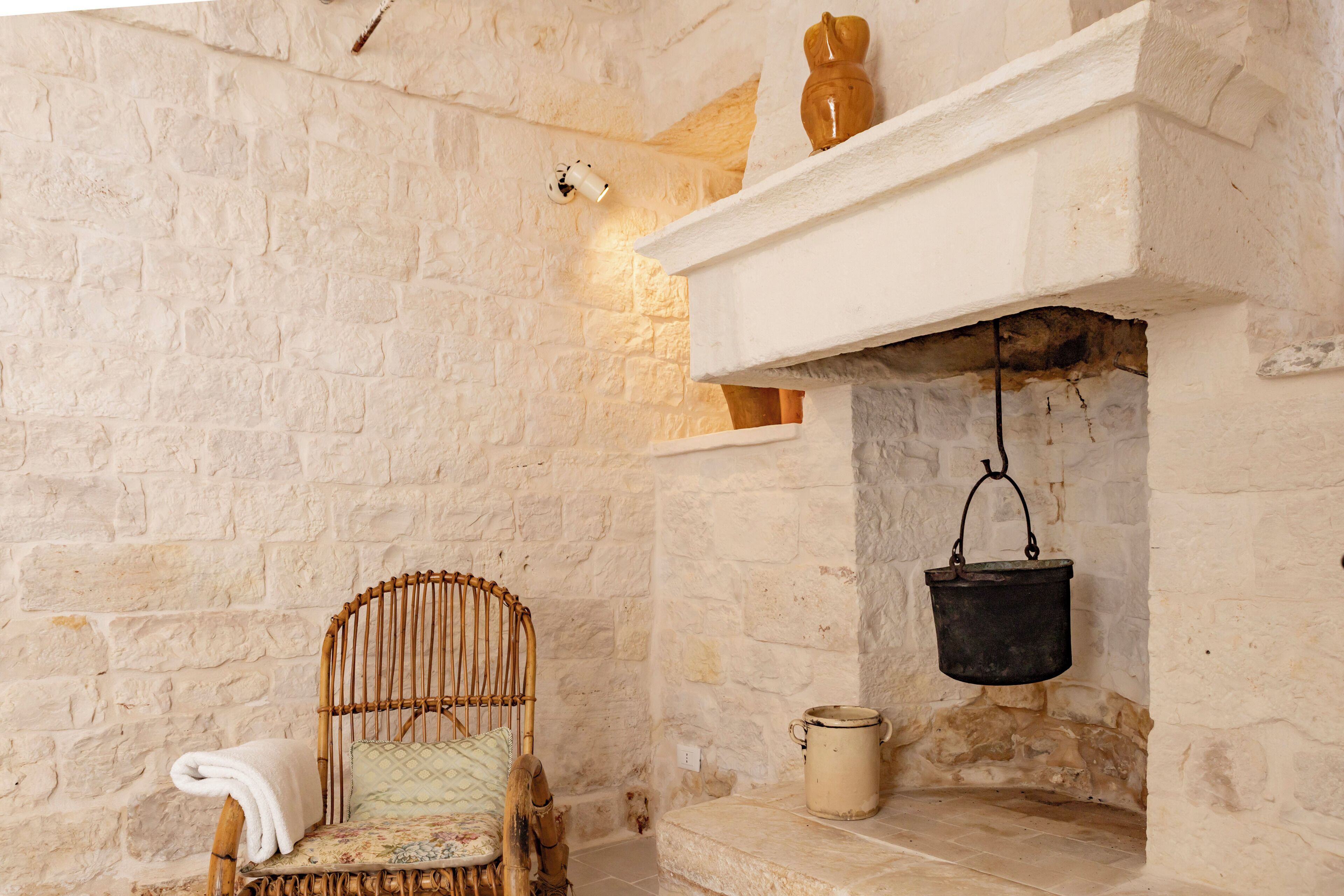 A rustic stone-walled room with a wicker chair, a white shelf with a vase, and a hanging pot.