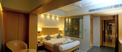 A modern hotel room with a large bed, a sitting area with a chair and a small table, and a glass-enclosed shower area.