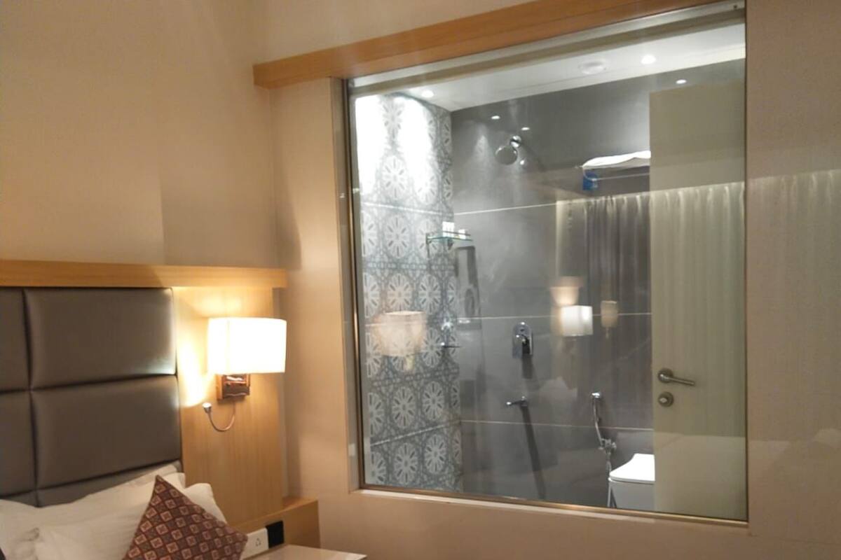 Una habitación de hotel con una cama grande, una mesita de noche con lámpara, una ventana con vistas al baño y un cabecero.