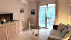 Un salon avec un canapé, un téléviseur, une petite table et une chaise. Il y a une fenêtre avec des rideaux et une vue sur l’extérieur.