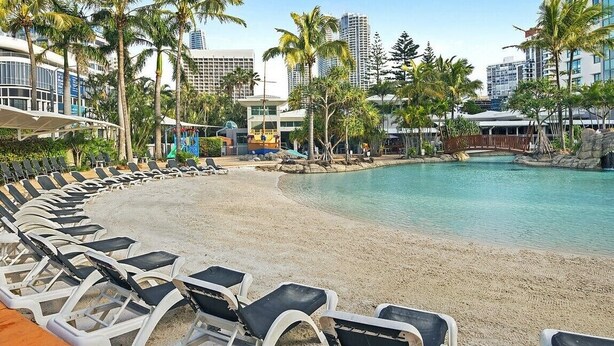 A resort pool area with lounge chairs, palm trees, and modern buildings in the background.