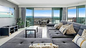 A modern living room with a gray sofa, a coffee table, and a TV on the wall. There is a balcony with a view of the city and a sailboat on the water.