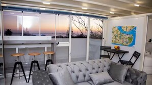 A modern living room with a grey tufted sofa, a bar area with stools, a dining table with chairs, and a large window with a view of the sunset. - TERRACE FLAT AT THE CENTER OF OLD CITY WITH JACUZZI (Istanbul)