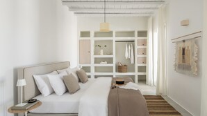 A modern bedroom with a bed, bedside table, lamp, and a view into a walk-in closet with shelves and hanging clothes. - Esti Villa Melissa - Executive Luxury Villa (Mykonos)