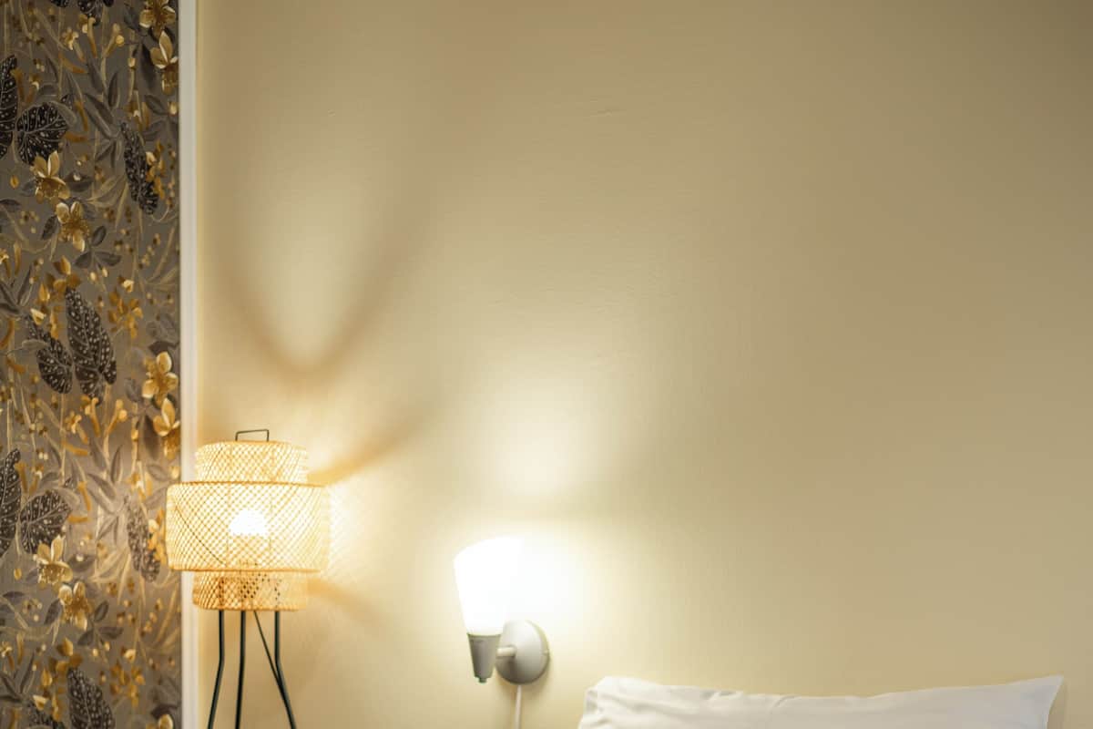 Un lit bien fait, avec du linge de lit blanc et un oreiller marron, une lampe fixée au mur et un papier peint à motifs floraux.