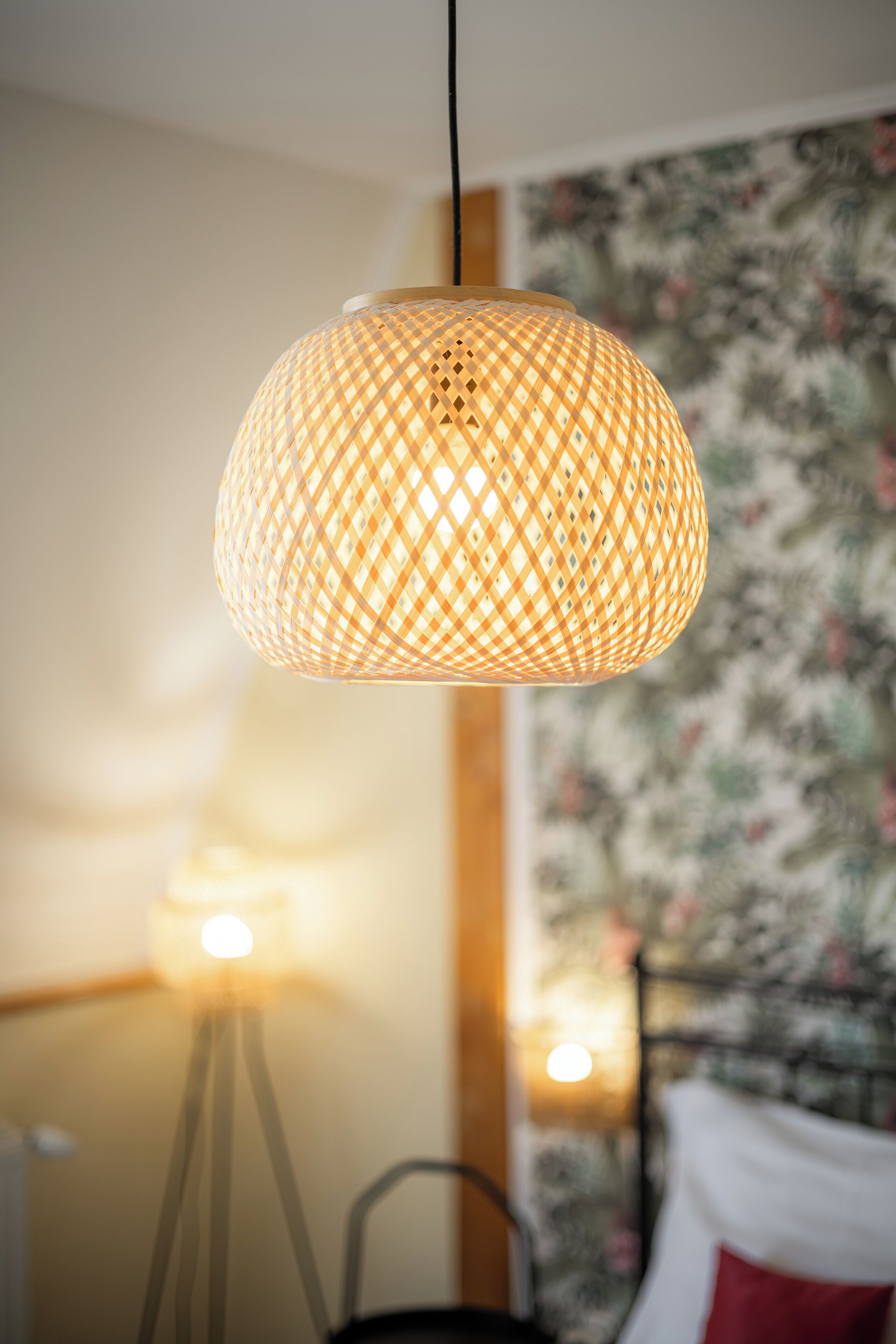A hanging lamp with a woven pattern, a bed with a red pillow, and a floral wallpaper background.