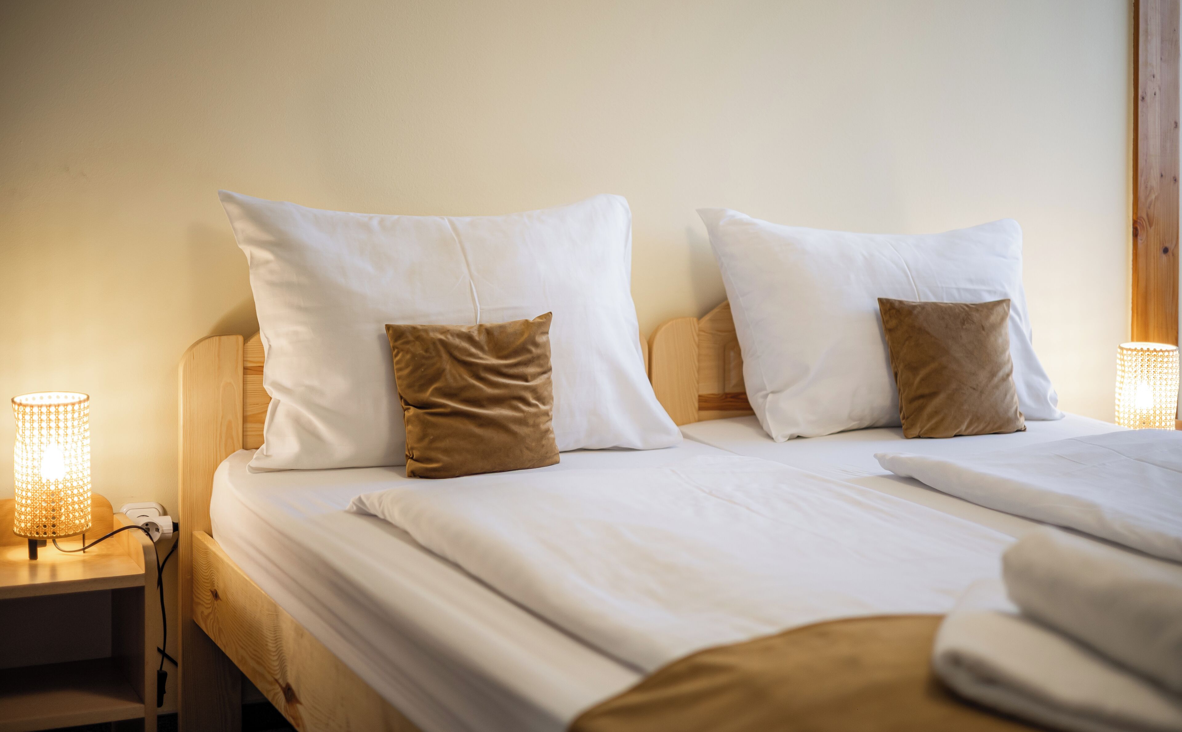 Una cama bien hecha con ropa de cama blanca y dos almohadas marrones, un cabecero de madera y una mesita de noche con una lámpara encendida.