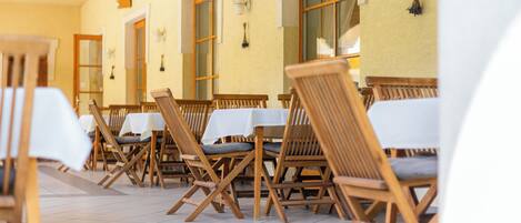 A row of wooden chairs with white tablecloths, arranged in a dining area with a tiled floor and beige walls.