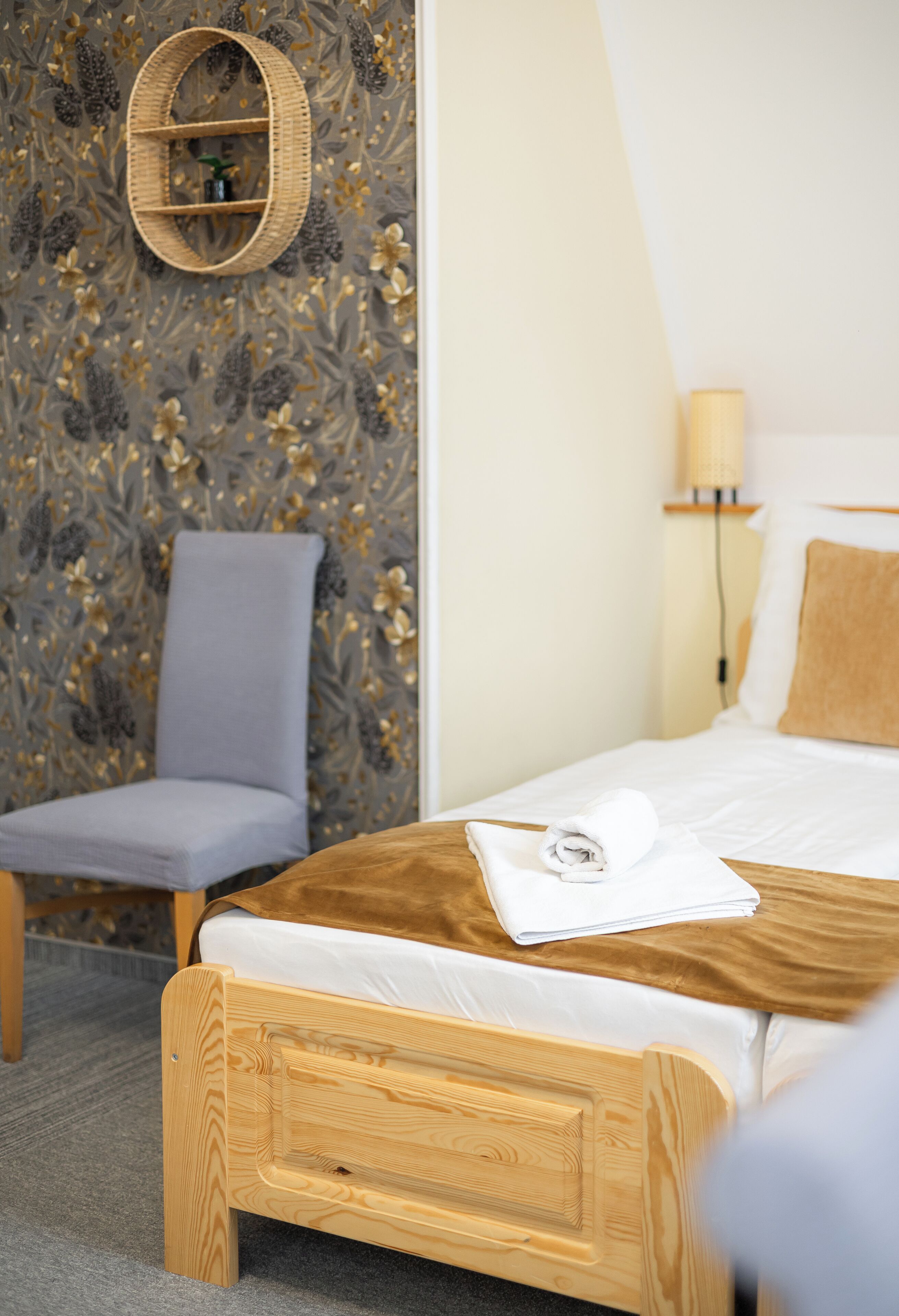 Een keurig opgemaakt bed met houten hoofdbord, een grijs gestoffeerde fauteuil, een wandplank van riet en een lamp op een nachtkastje.
