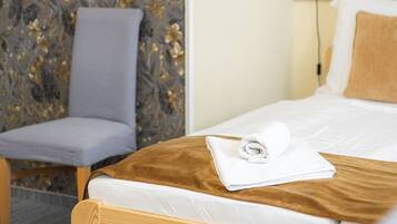 Een keurig opgemaakt bed met houten hoofdbord, een grijs gestoffeerde fauteuil, een wandplank van riet en een lamp op een nachtkastje.