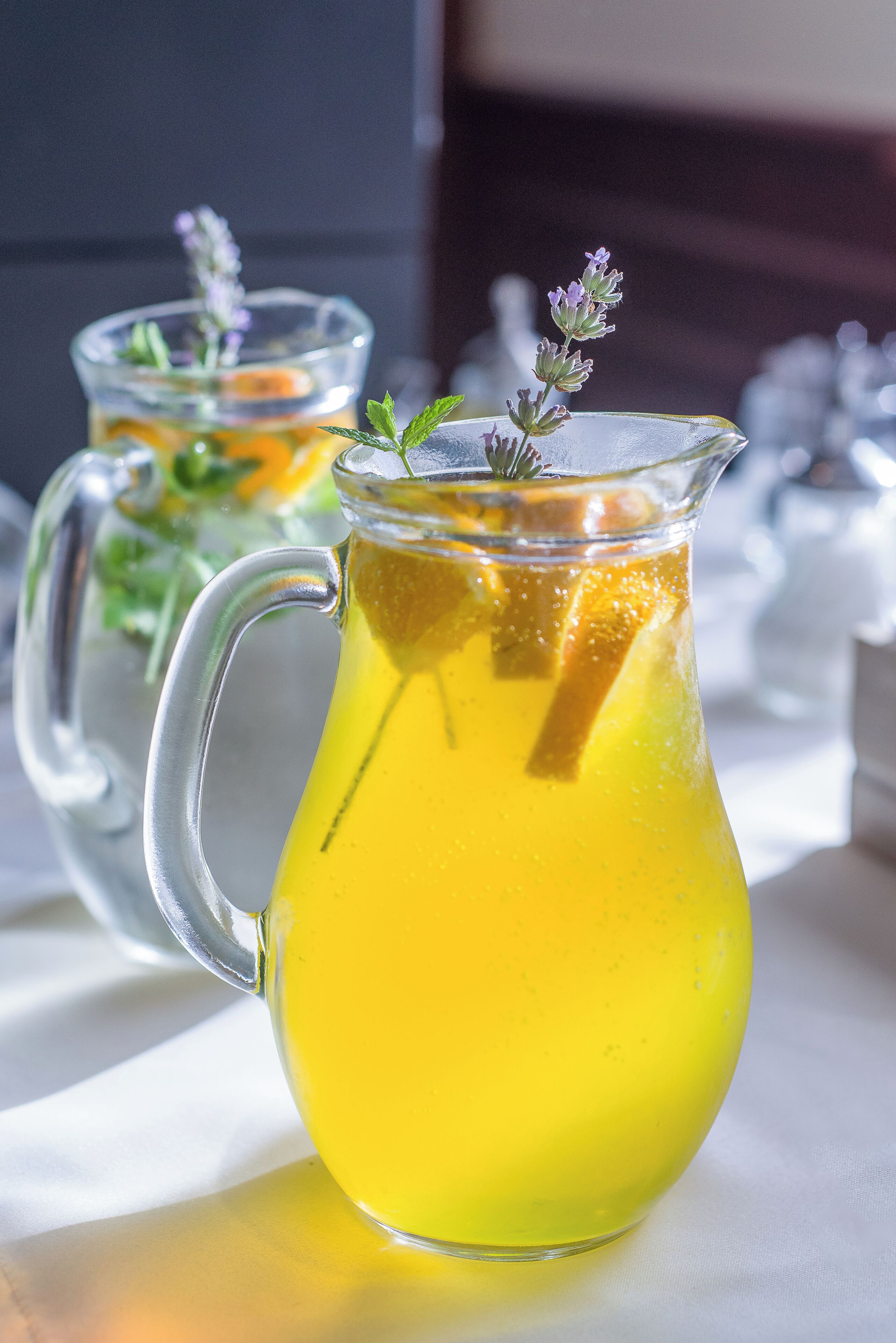 A pitcher of yellow beverage with a handle, garnished with a sprig of lavender, on a white surface.