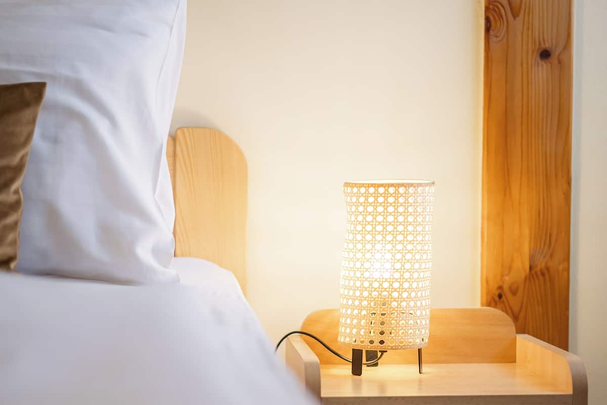 Un lit bien fait, avec du linge de lit blanc et un seul oreiller marron, une table de chevet en bois surmontée d’une lampe en osier et un panneau mural en bois.