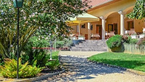 A hotel entrance with a covered patio, steps leading to the entrance, and a garden area with a pathway. - Regia Panzió és Étterem (Halászi)