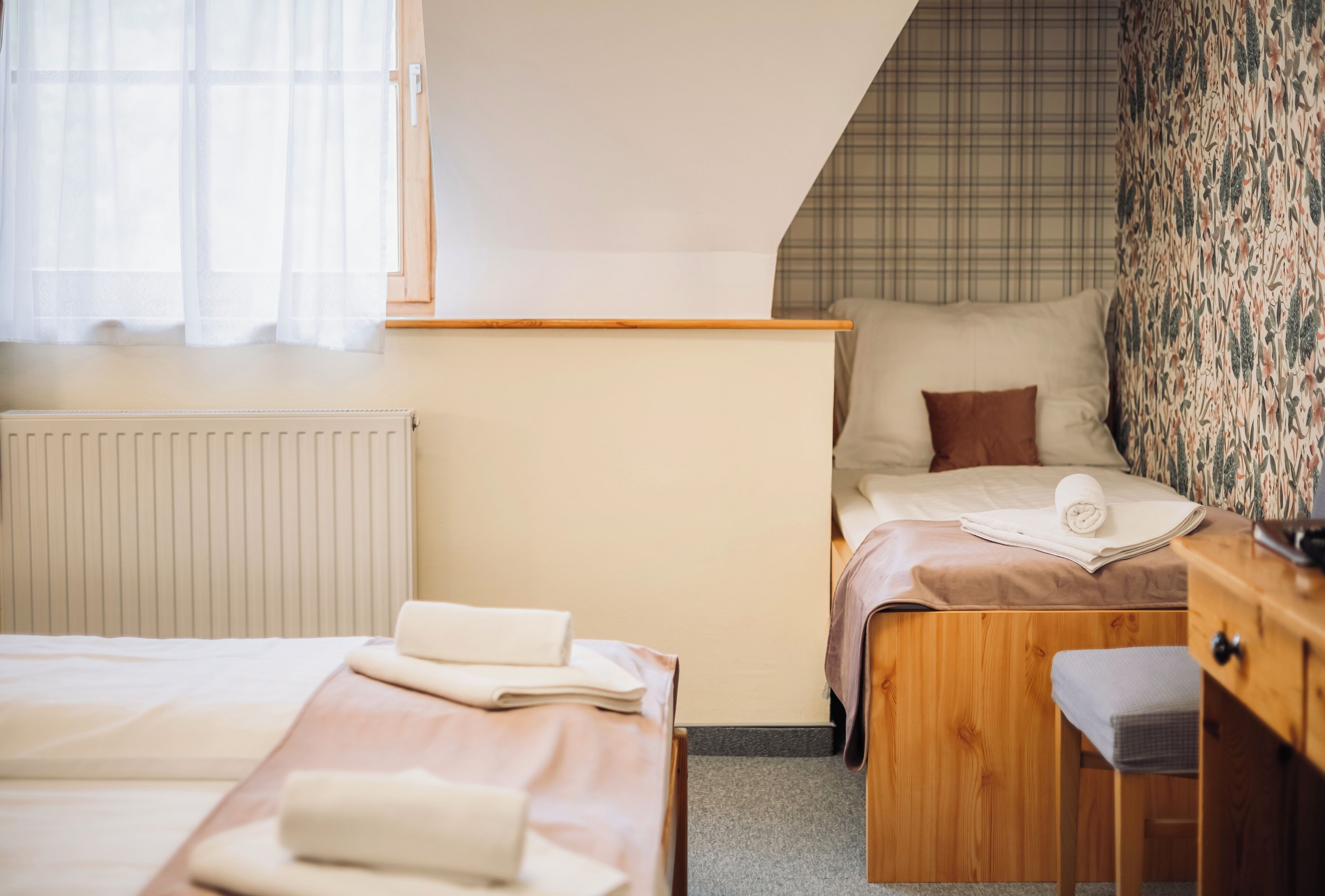 A hotel room with two beds, a wooden desk, a radiator, and a window with sheer curtains.
