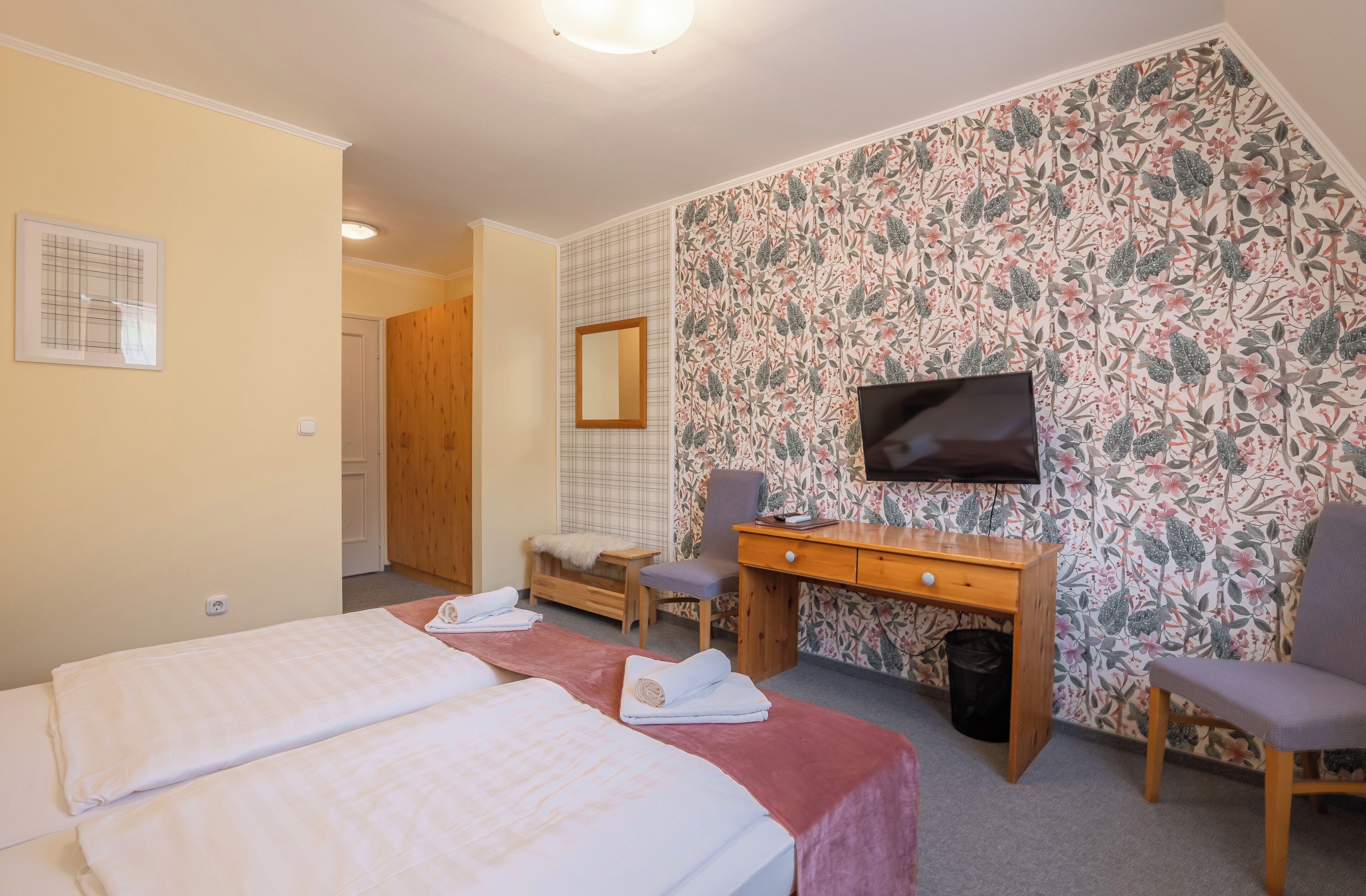 Ein Hotelzimmer mit zwei Betten, einem Schreibtisch, einem Fernseher und blumigem Wandpapier.