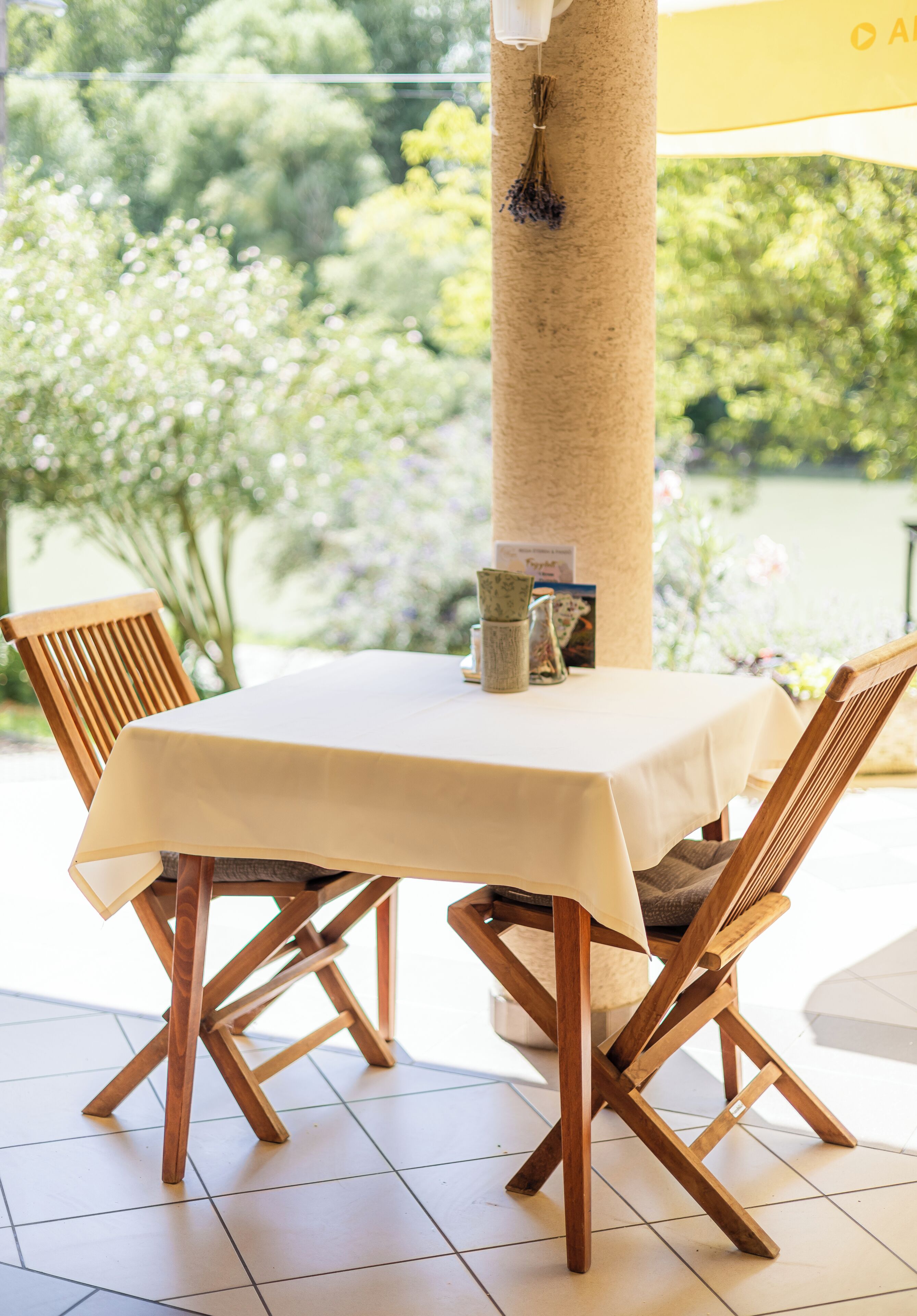 Una mesa con mantel blanco, dos sillas de madera, un florero y un menú en un patio soleado con vistas a árboles y una masa de agua.