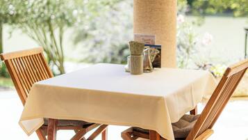 Una mesa con mantel blanco, dos sillas de madera, un florero y un menú en un patio soleado con vistas a árboles y una masa de agua.